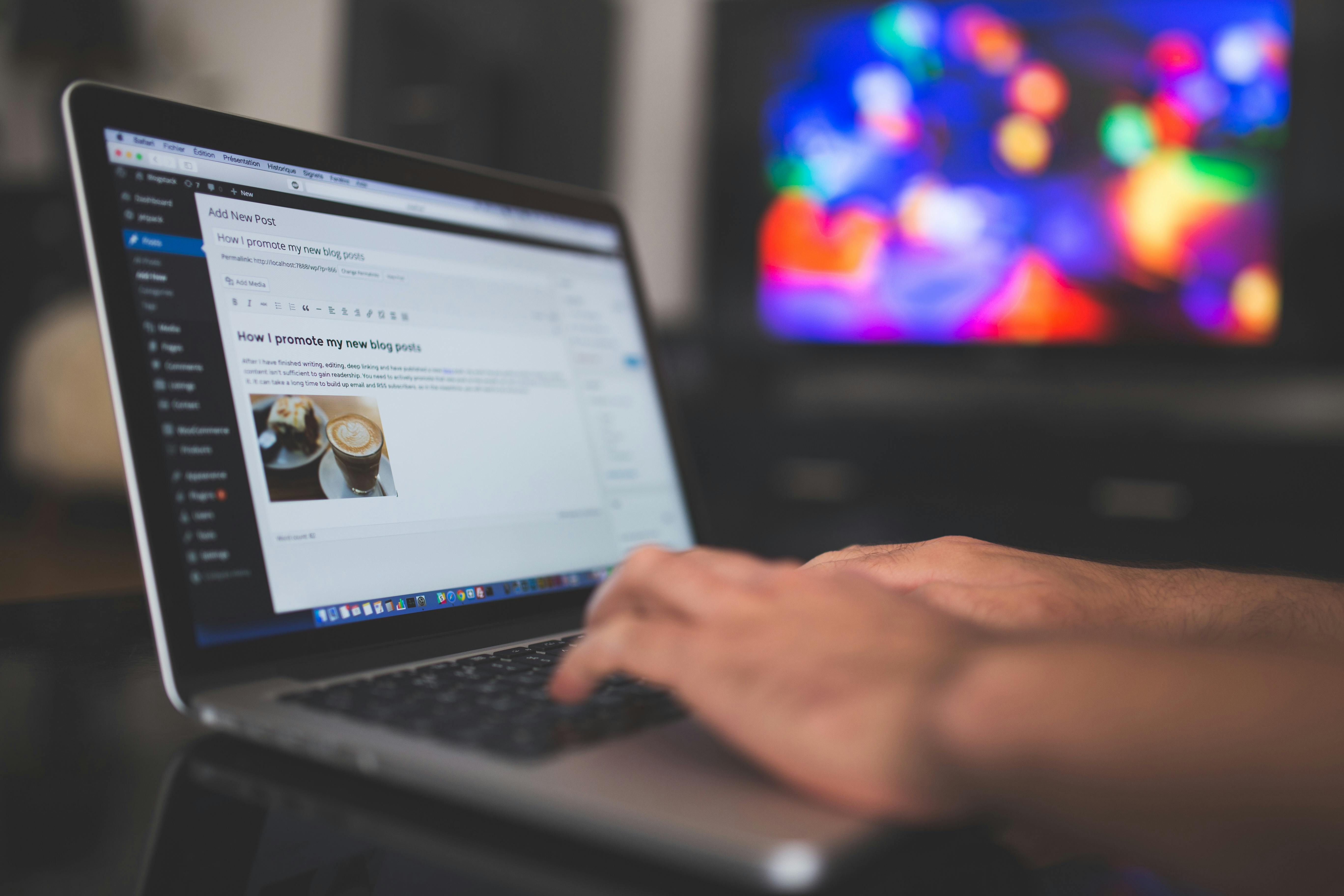 Person's hand on laptop keyboard displaying WordPress blog post editor with title "How I promote my new blog posts" and coffee cup image, with blurred colorful lights visible on monitor in background.